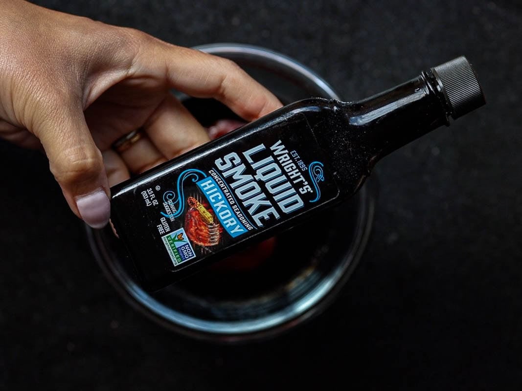 Hand holding a bottle of Wrights Liquid Smoke with Hickory flavor above a clear glass bowl on a dark surface. The label features cooked meat and the bottle is dark with a black cap.